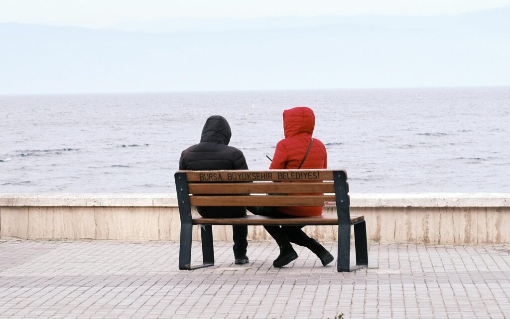 Two individuals in jackets sit on a bench by the sea in Bursa, Turkey, with a quiet atmosphere.